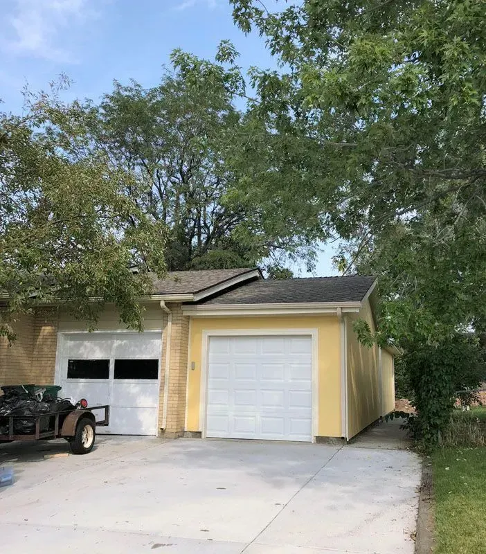 Two-car garage with white doors, yellow exterior, and concrete driveway; surrounded by trees.