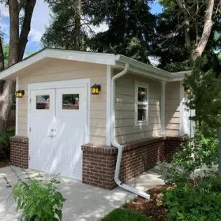 Tan and brick garage with white doors and window, surrounded by trees and greenery.