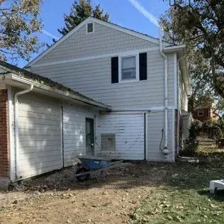 Side view of a two-story house with light siding and dark shutters, next to a garage.