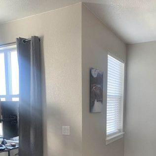 Corner of a room with a window, blinds, and a painting; neutral-toned walls and a curtain.