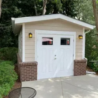 Tan-sided garage with white door, brick accents, and two sconce lights, set in a yard with greenery and a concrete driveway.