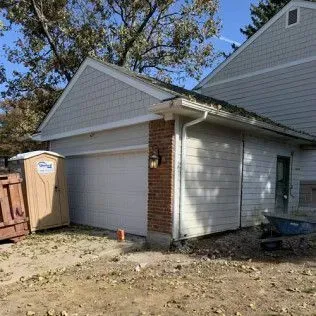 Garage with white door, tan siding, and brick accent. A portable toilet sits nearby.