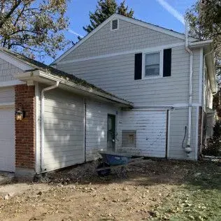 Side view of two-story house with light siding, black shutters, brick and dirt yard.