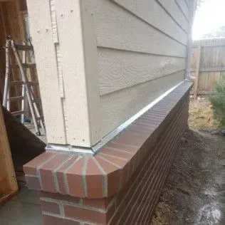 Brick wall supporting a beige siding corner, with a gutter and ladder in the background.