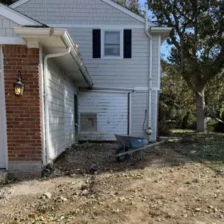 Side view of a house with brick and gray siding. A wheelbarrow sits on dirt ground next to the house.
