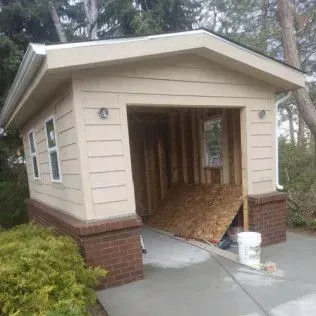Tan-colored garage under construction with exposed interior frame and a partially open garage door; bricks at base.