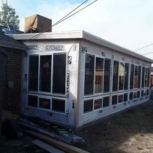 Sunroom under construction with many windows and white framing attached to a brick house.