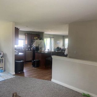 Open-concept living space with kitchen visible; light-colored walls, wood floors, and gray carpet.