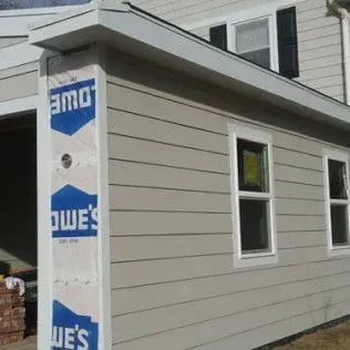 Exterior of a building with light gray siding, white trim, and two windows. A garage and Lowe's wrap are visible.