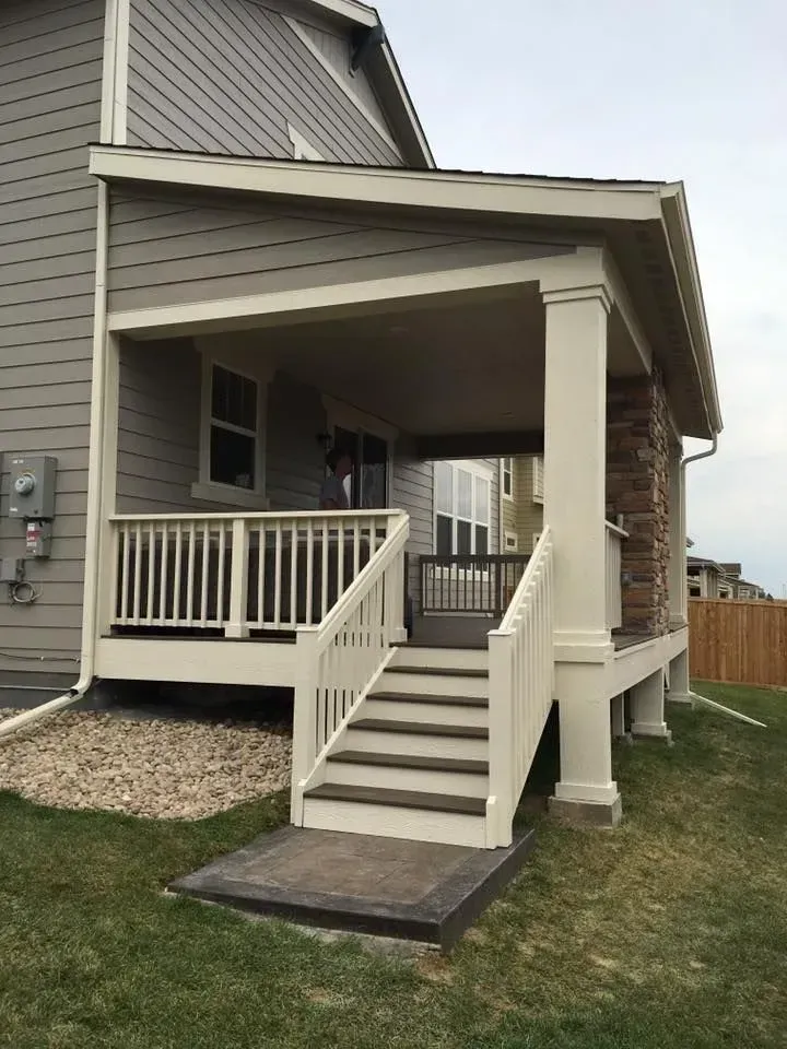 Beige porch with steps and railing attached to a gray house, with grass in the foreground.