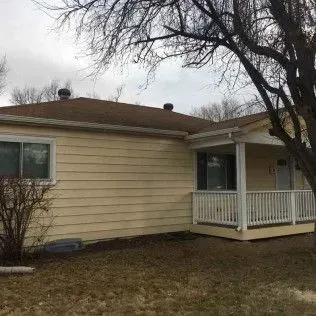 Yellow house with a porch and brown roof, bare tree in the foreground. Overcast sky.