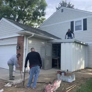 Men working on the side of a house. One is on the roof of a small structure, others are on the ground near the structure.