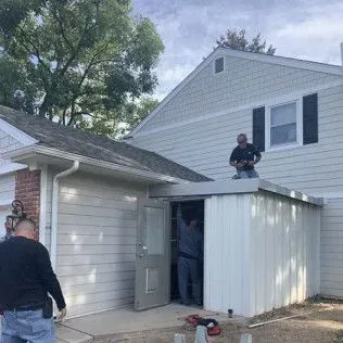 Workers installing siding on a shed attached to a house. One man is on the shed roof.