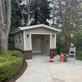 Small detached garage with tan siding, brick accents, and open doorway on a concrete driveway.