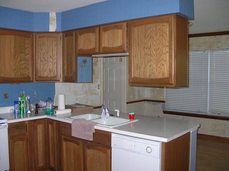 Kitchen with light wood cabinets, white countertops, blue patterned wallpaper, and a window with blinds.