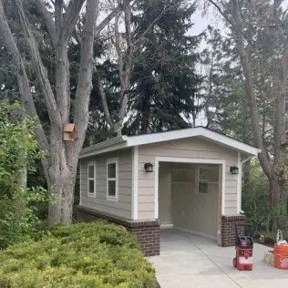 Tan garage with open entrance, brick base, and windows. Driveway with greenery and trees.