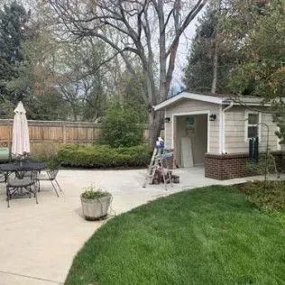 Backyard patio with garage, trees, green lawn, and a closed umbrella.