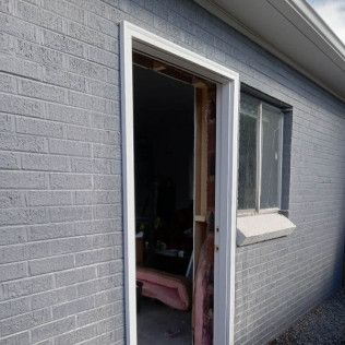 Exterior shot of a gray painted brick building with a white door frame and open doorway. A small window sits to the side.