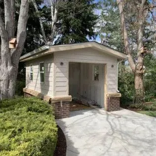Small tan garage with brick pillars; trees and greenery surround.