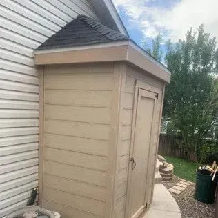 Tan shed with a black roof next to a house with white siding and a green yard.