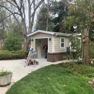 Garage under construction with tan siding and brick base, near trees and lawn.