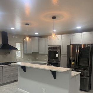 Modern kitchen with light gray cabinets, black range hood, stainless steel appliances, and island with pendant lights.