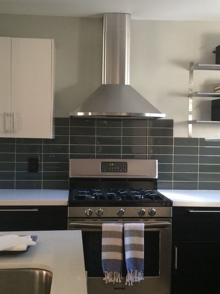 Kitchen with stainless steel range, hood, and dark tiled backsplash. White cabinets, countertops, and shelving.