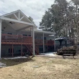 House with wooden decks, gazebo, and yard. Snow patches on ground, trees in background. Cloudy sky.