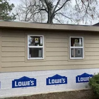 Exterior of a house with two windows and beige siding, Lowe's branded wrap, and bare trees in background.