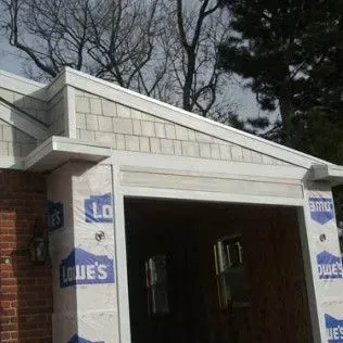 Garage under construction with shingled gable, white trim, and brick base; covered in Lowe's wrap.