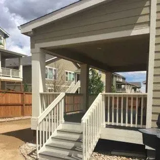 Covered porch with stairs, railing, and tan siding. Overcast sky in background.