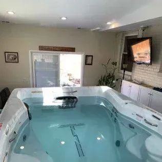 Indoor swim spa filled with clear water, near a sliding door, television on wall.