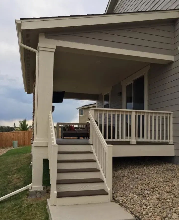Beige and gray porch with steps, railing, and overhanging roof. Brown steps lead to seating area.