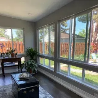 Sunroom with large windows overlooking a yard and a wooden fence. Includes plants, a table, and a trunk.