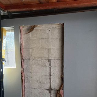 Opening in a gray wall reveals a cinder block wall. Framing with insulation is exposed around the opening.