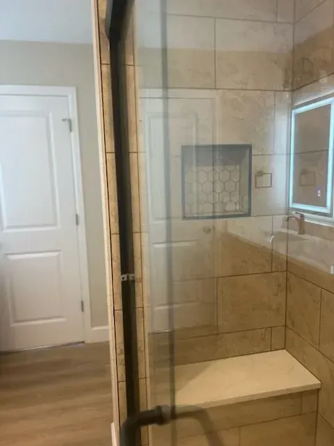 Shower stall with glass door, bench seat, beige tiles, and a white door in the background.