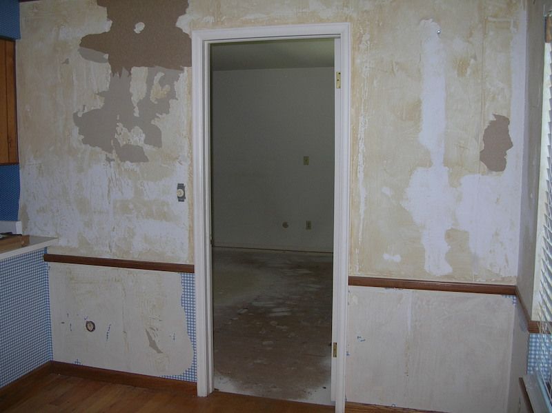 Interior view of a room with partially stripped wallpaper. Doorway leads to an adjacent room.