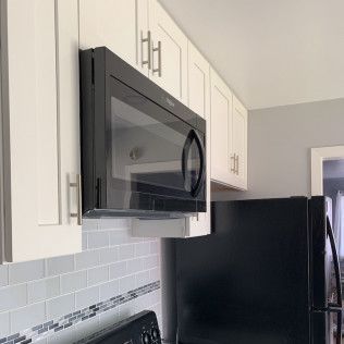 Black microwave over a stove with cabinets in a kitchen. Black refrigerator to the right.