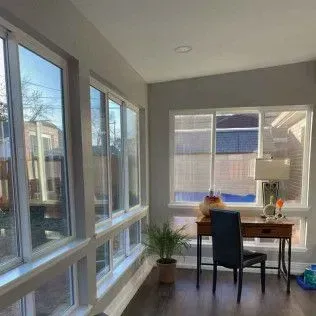Sunroom with many windows, desk, chair, and a plant. Brown floor, gray walls, and bright sunlight.