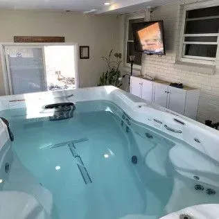 Indoor swim spa filled with clear water, facing a brick wall with a mounted TV and white cabinets.