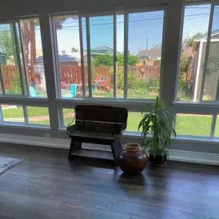 A room with dark wood floors, large windows overlooking a backyard, bench, pot, and plant.