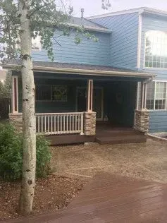 Blue house with porch and stone-columned supports. Brown deck, white railing, tree in foreground.