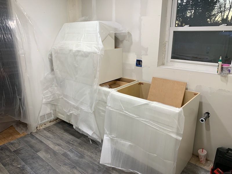 Kitchen cabinets, wrapped in plastic, under construction near a window.  Gray flooring and white walls.
