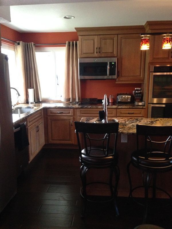 Kitchen with medium-toned wooden cabinets, dark floors, and red-orange wall.  Bar stools sit at the island.