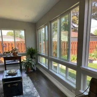 Sunroom with large windows, dark wood floor, plants, a table, and a trunk.  Bright daylight.