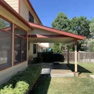 Tan house with a red-trimmed awning over a concrete patio. Green bushes and grass surround the outdoor space.