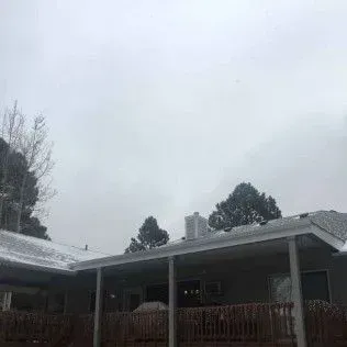 Snow-covered house with a wooden deck and trees against a cloudy gray sky.