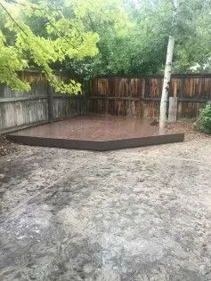Brown, triangular outdoor concrete patio with a wooden border, in a backyard setting.