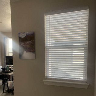 Window with white blinds, partially open, letting in light. A painting hangs on the wall next to it.