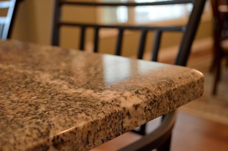 Close-up of a granite countertop's edge with a reflective surface. A black chair frame is nearby.